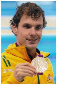 Paralympian Matt Levy holding a gold medal. He is smiling and is wearing a green and gold jacket. Swimming lanes appear in the background of the image.