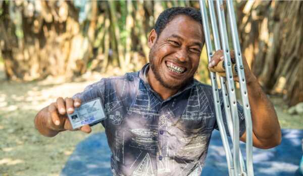 A man, wearing a blue short sleeved shirt is holding crutches with one hand and is smiling.